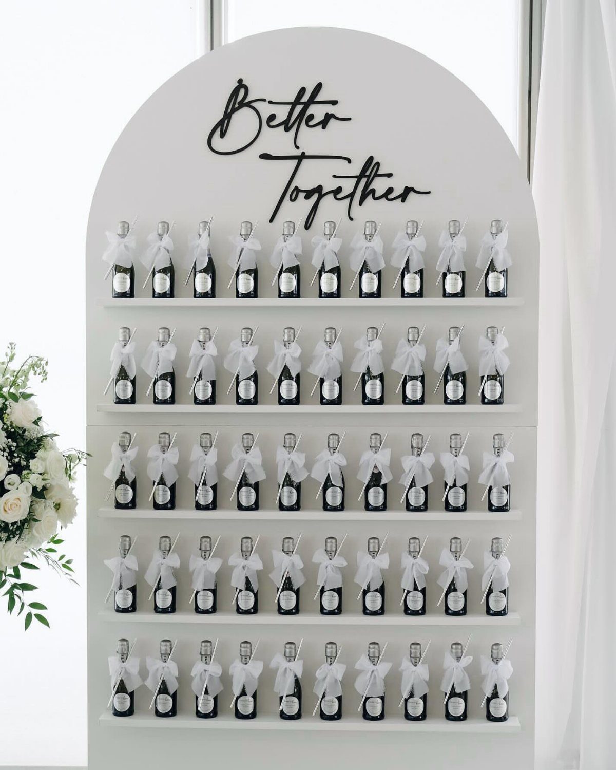 Mini champagne bottles with white ribbons on a white arched display shelf