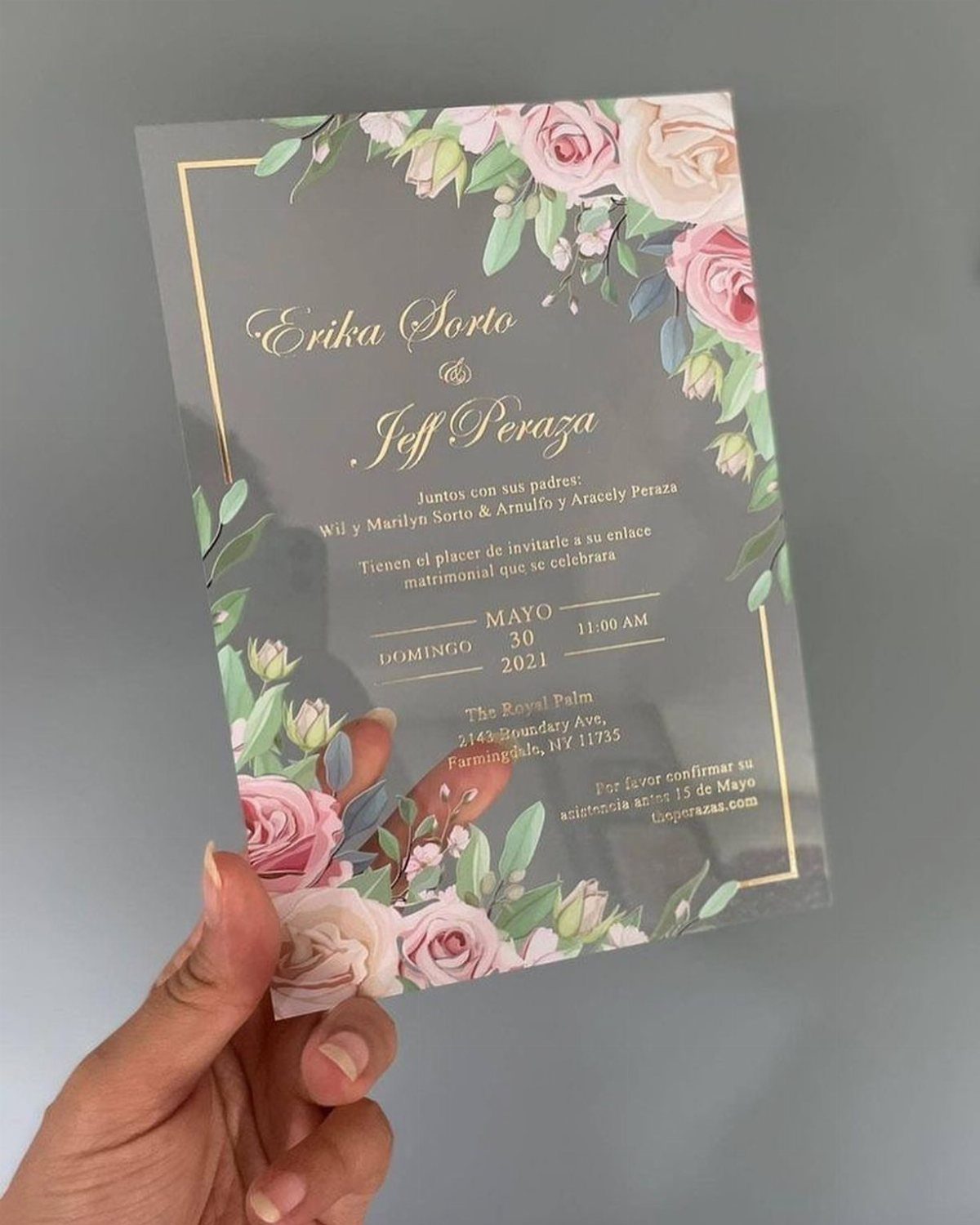 Transparent acrylic wedding invitation with gold calligraphy and pink floral corner accents.