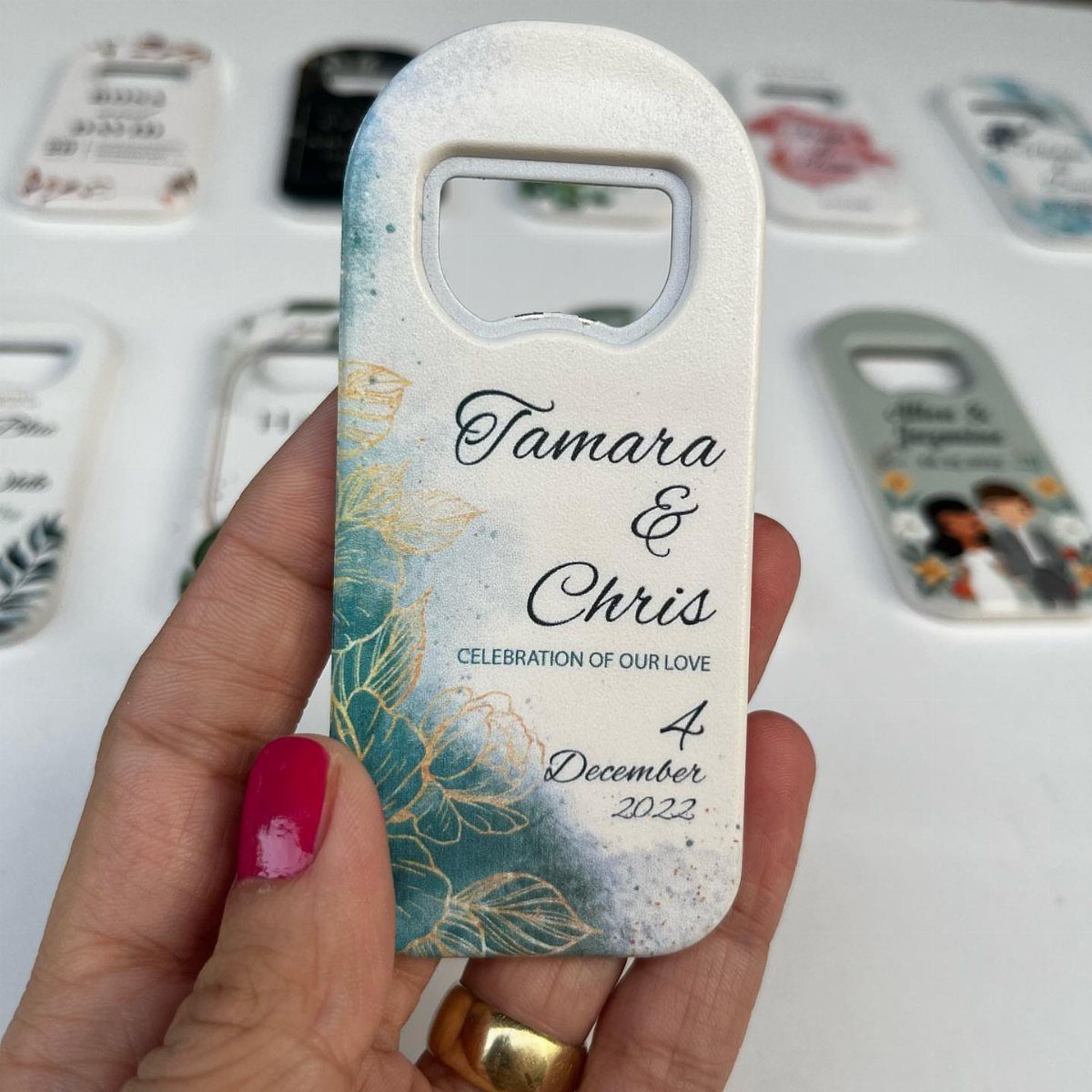 Personalized white bottle opener with floral design and wedding names held by hand.