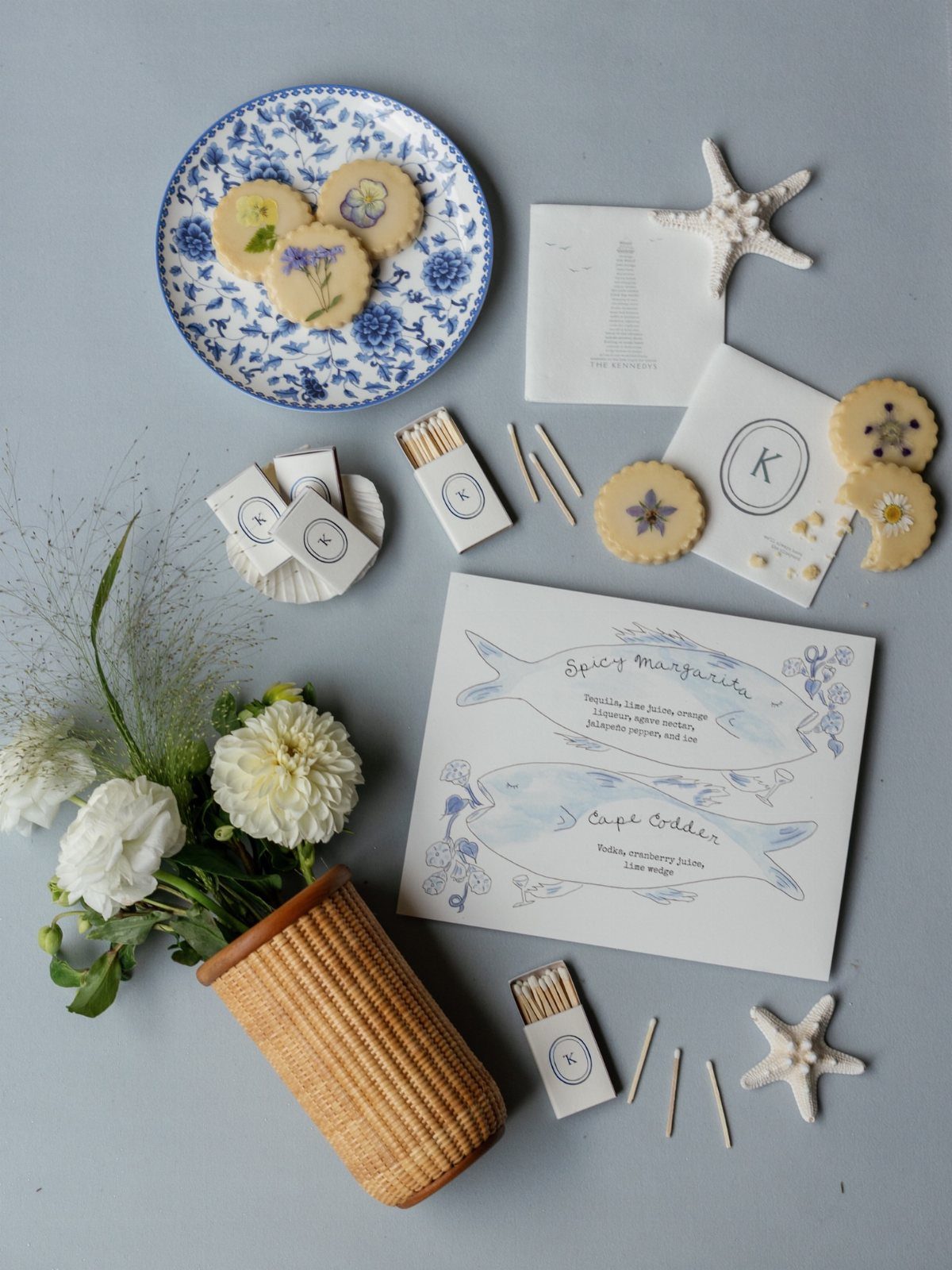 Customized white matchboxes with blue logos arranged with floral cookies and cocktail menus.