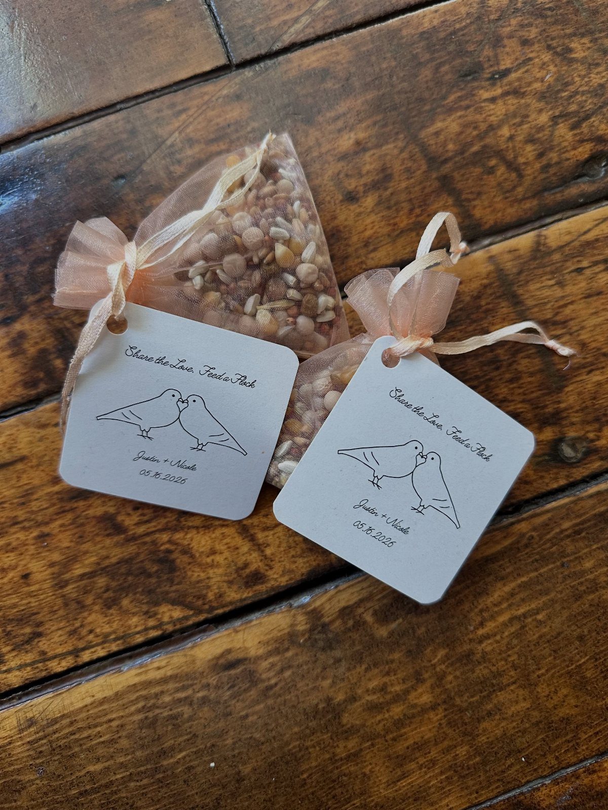 Two birdseed favor packets with peach ribbons and bird illustration tags on wood.
