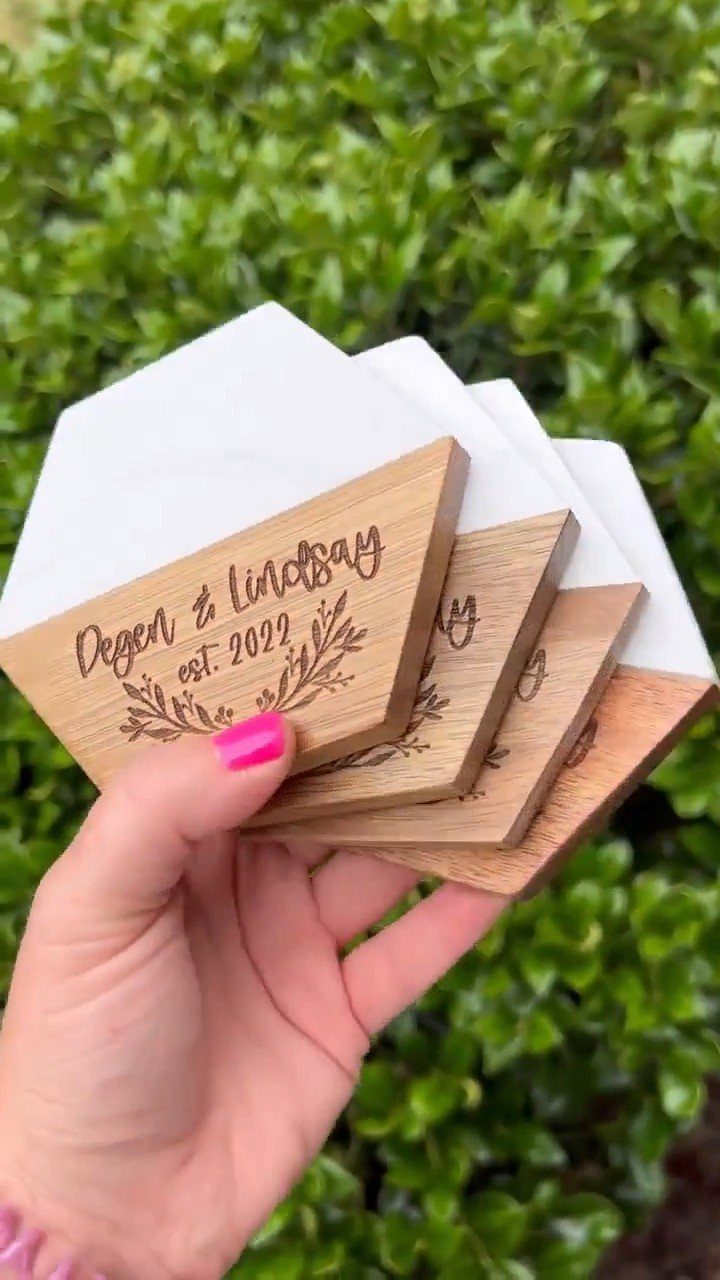 Hexagonal wooden coasters engraved with names and a date held against green leaves.