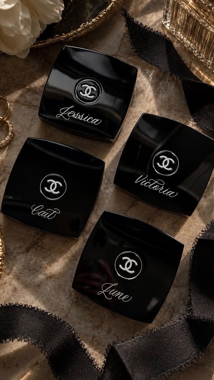 Four black square hand cream jars with personalized names on a marble surface.