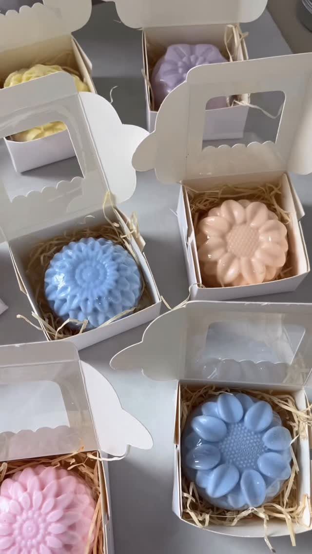 Flower-shaped pastel soaps in white gift boxes with brown shredded paper filler.