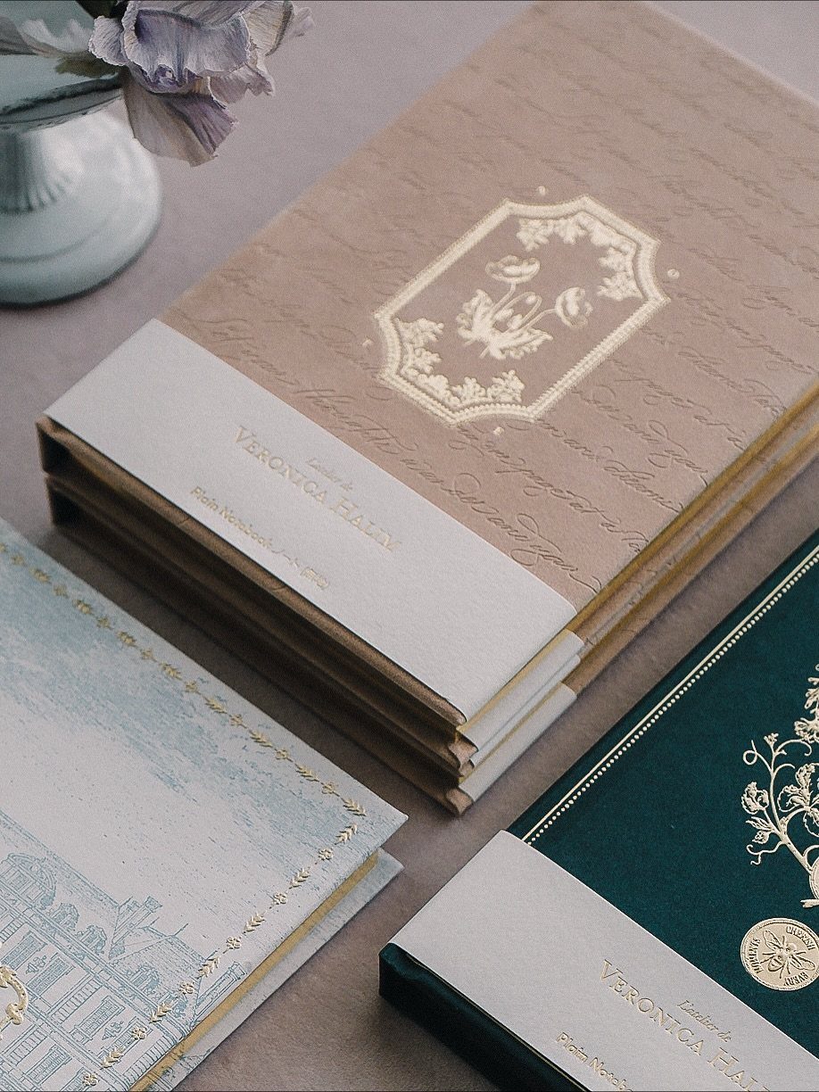 Personalized hardcover journals in beige, blue, and green with gold foil embossed details.