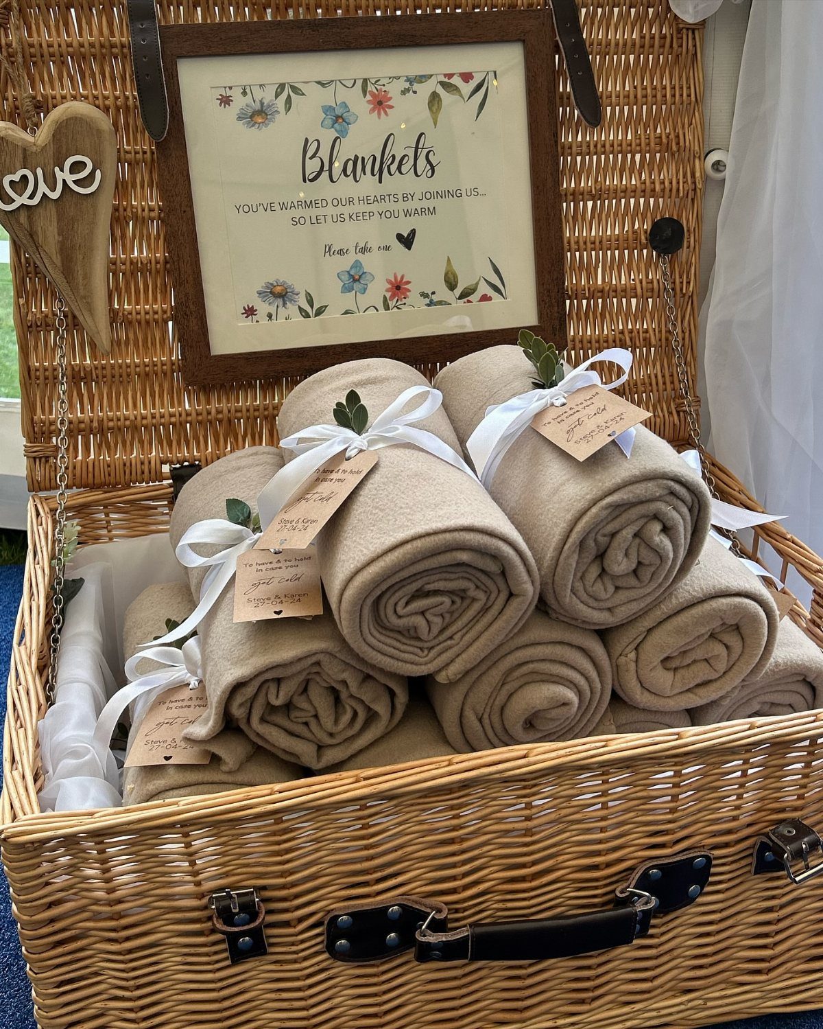 Beige rolled throw blankets with white ribbons in a wicker basket with sign.