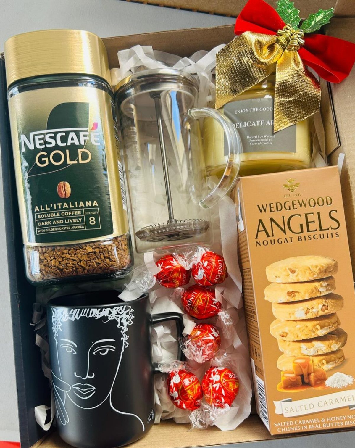 Gift box with coffee, French press, red-wrapped chocolates, biscuits, and a black mug.