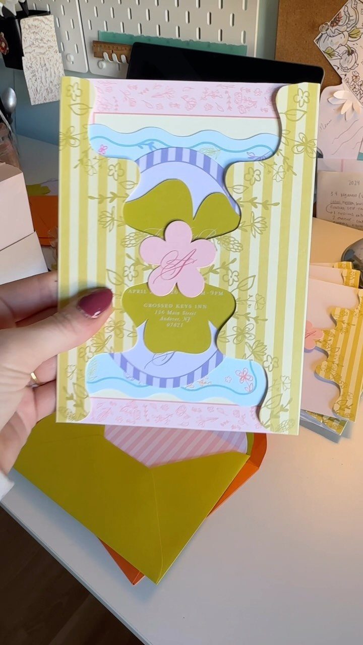 Layered colorful wedding invitation with yellow stripes and pink floral cutouts held by hand.