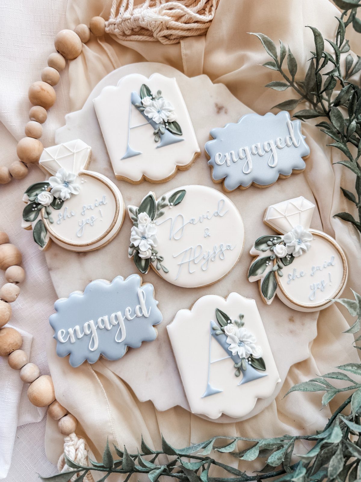 Decorated engagement cookies with rings and initials on a marble slab with greenery.