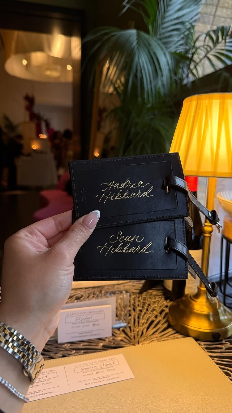 Two black leather luggage tags with gold cursive names held by a hand.