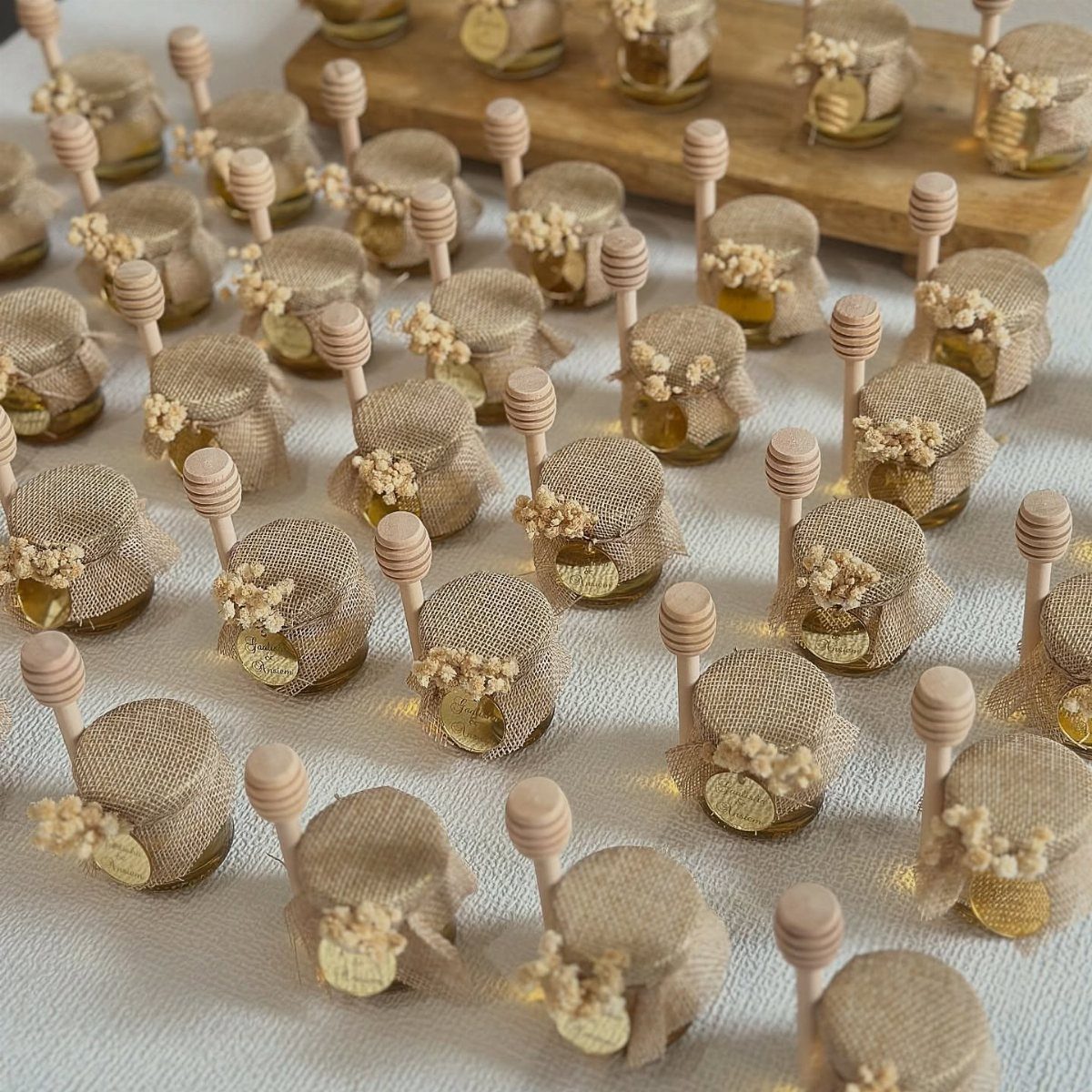 Mini honey jars with burlap lids and wooden dippers arranged on white fabric.