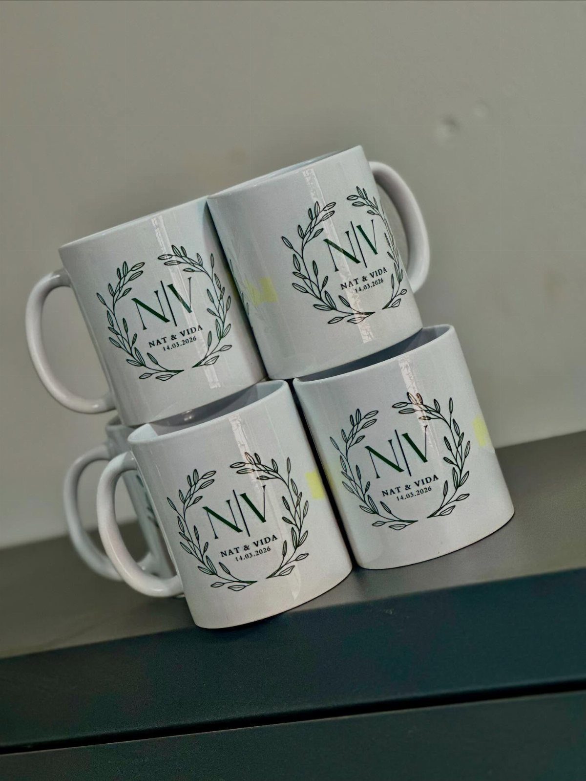 Four white ceramic mugs with personalized black wedding logos and names stacked together.