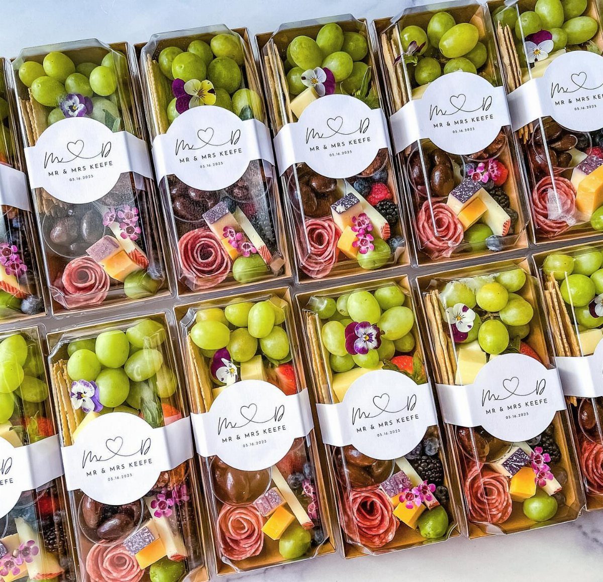 Charcuterie snack packs in clear plastic boxes with white ribbons and personalized labels.