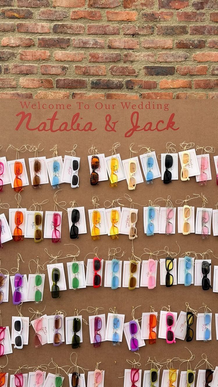 Colorful sunglasses hanging on a wedding welcome sign against a brick wall.