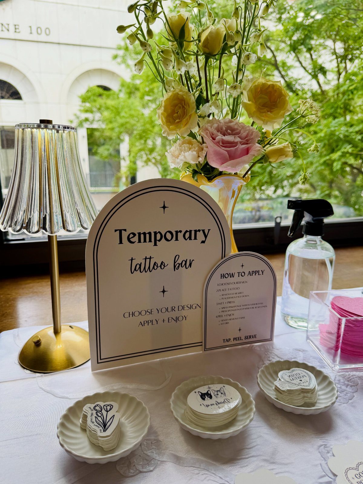 Temporary tattoo bar with designs on small plates, a sign, and floral arrangements.