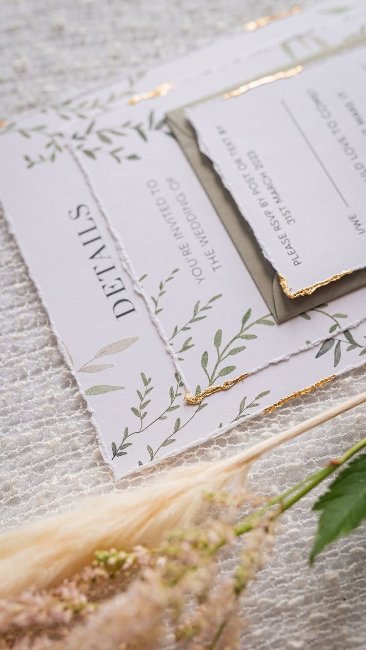 Wedding invitations with gold deckled edges and green botanical leaf illustrations on white paper.