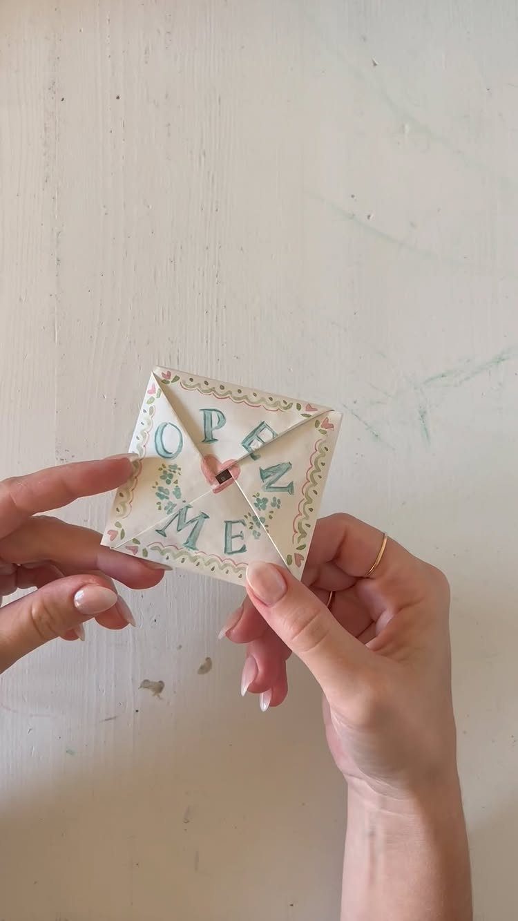 Hands holding a square folded paper invitation with floral borders and blue text.