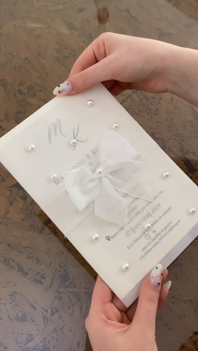 White wedding invitation with a fabric bow and pearl accents held by hands.