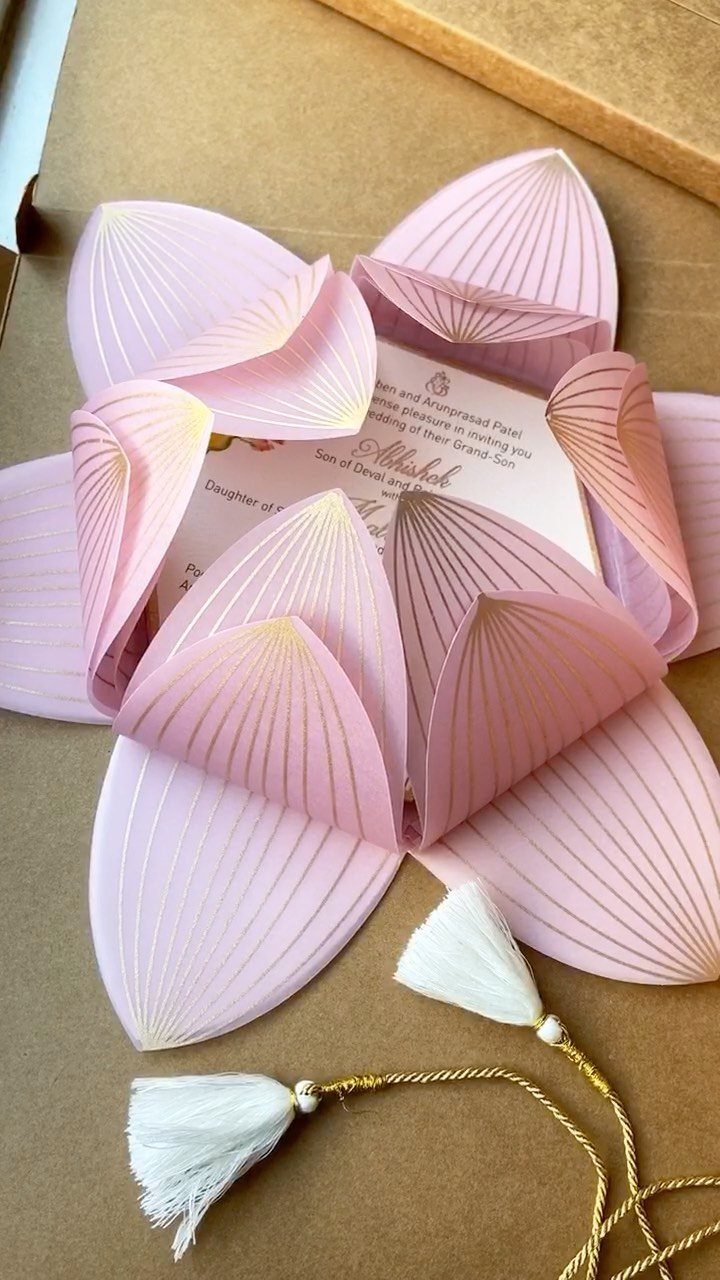 Pink paper invitation folded into a lotus flower with gold accents and white tassels.