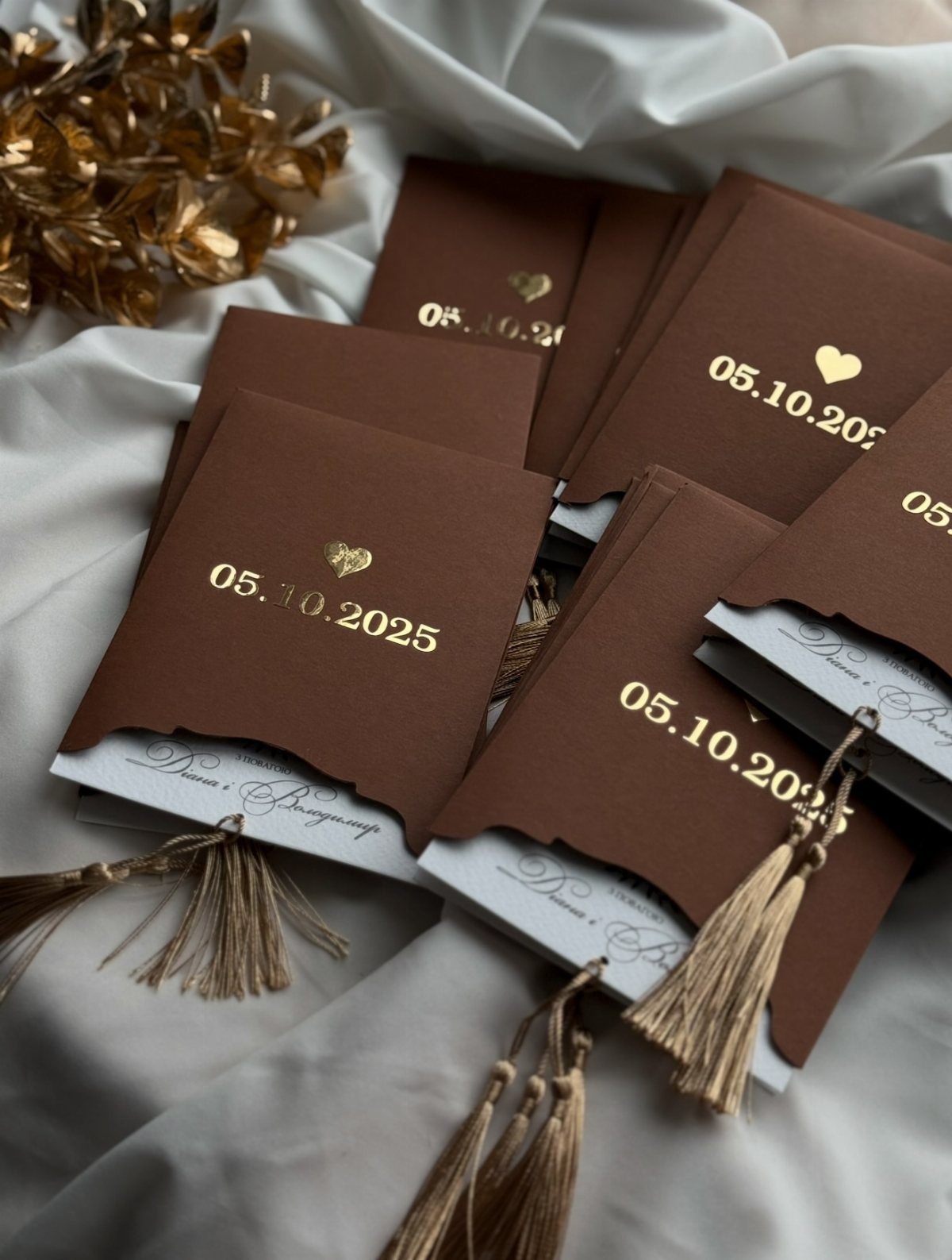 Brown wedding invitations with gold foil lettering and tassels on white fabric.