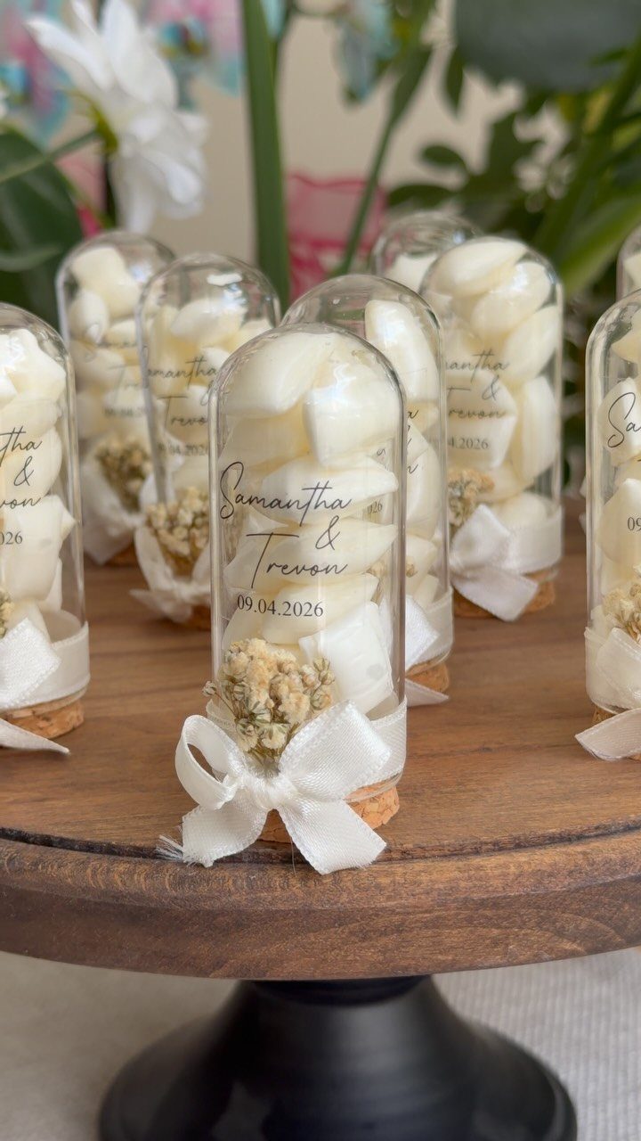 Glass vials with white petals and ribbons used as wedding invitations on wood.