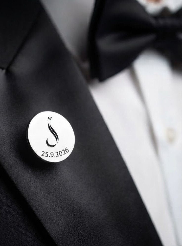 Silver lapel pin with a custom emblem and date on a black tuxedo.