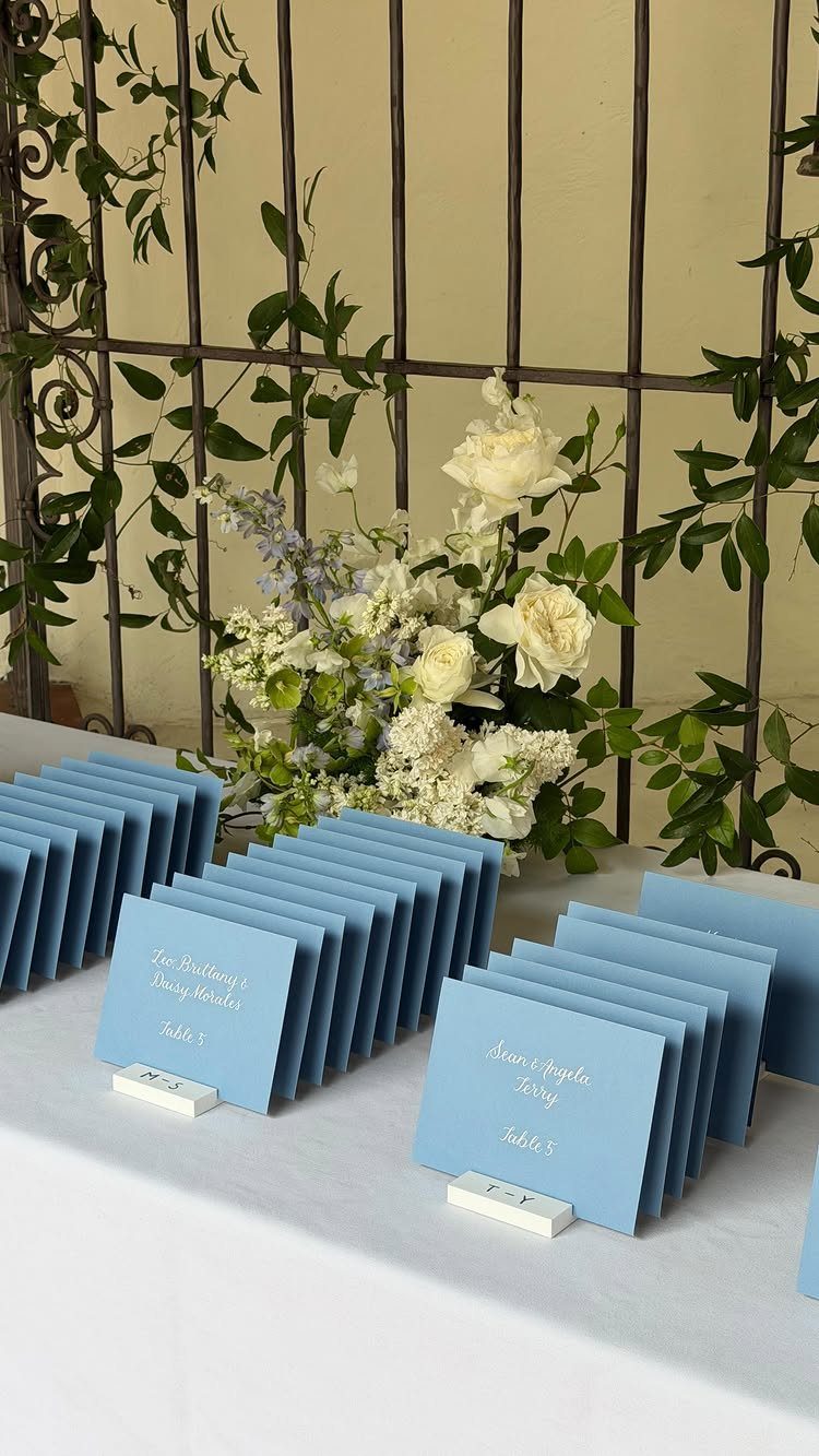 Blue wedding place cards arranged on a white table with white floral arrangements.