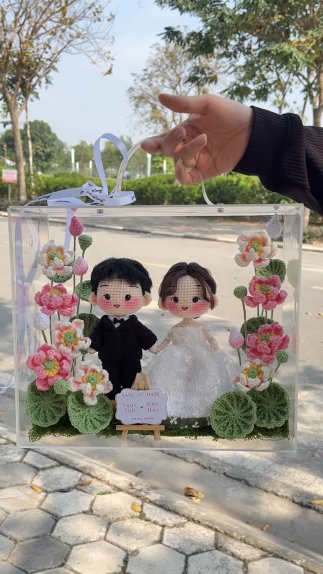 Crochet amigurumi bride and groom dolls inside a clear decorative floral display box.