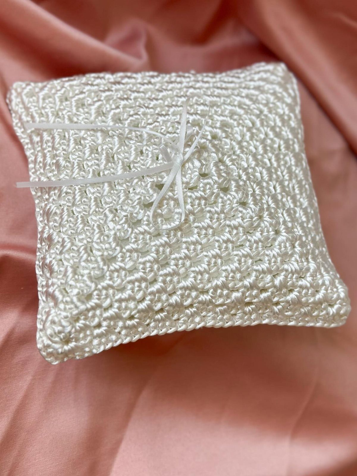 Small white crocheted pillow with a ribbon bow on pink satin fabric