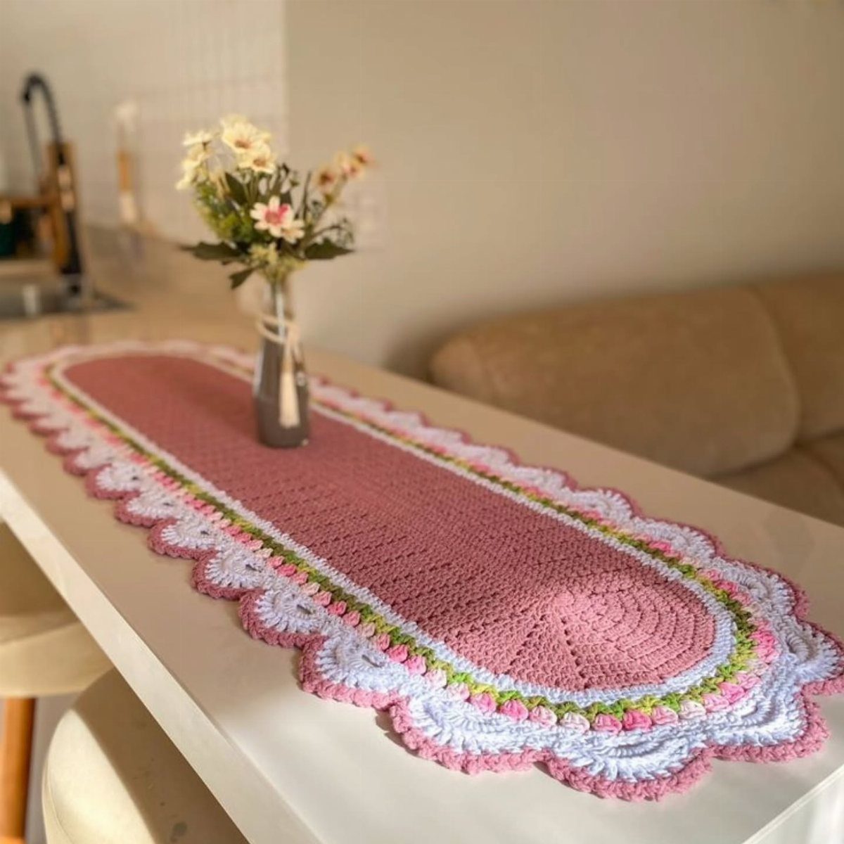 Pink crochet table runner with white scalloped edges and a vase of flowers.