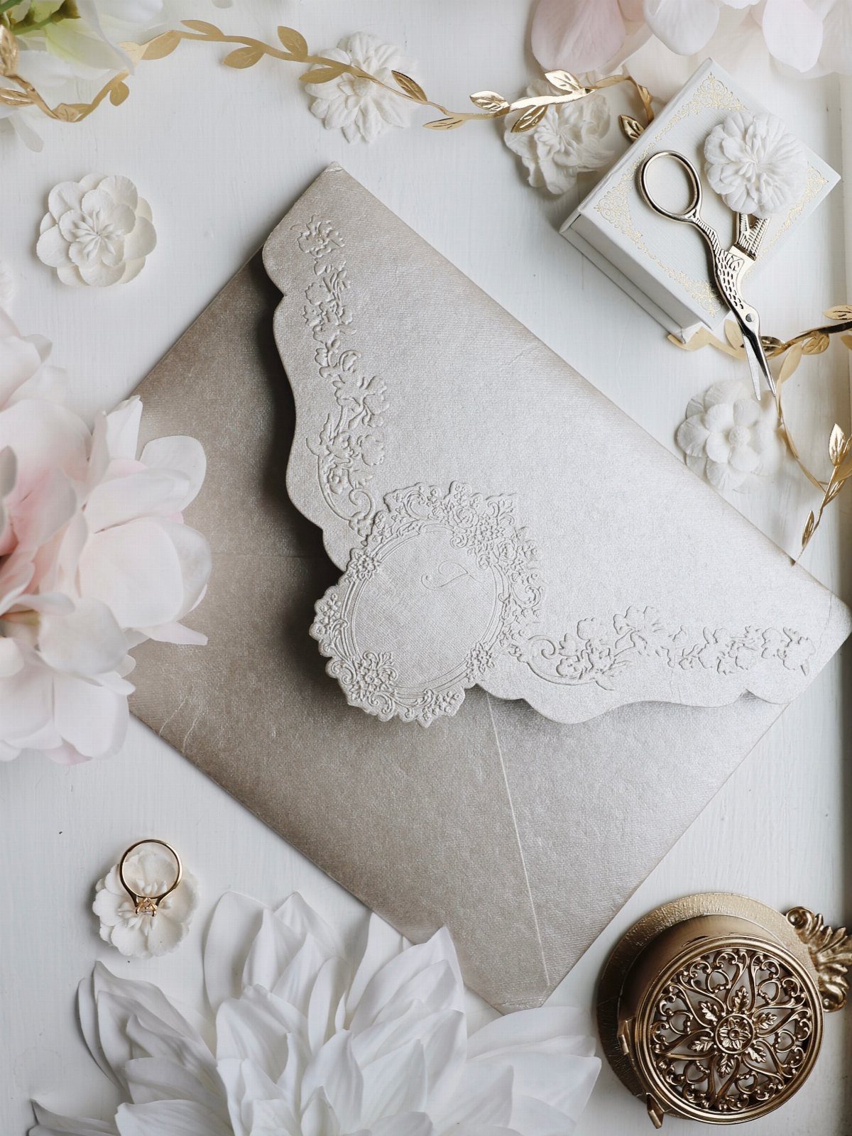 Embossed white wedding invitation with floral patterns on a taupe envelope and flowers.