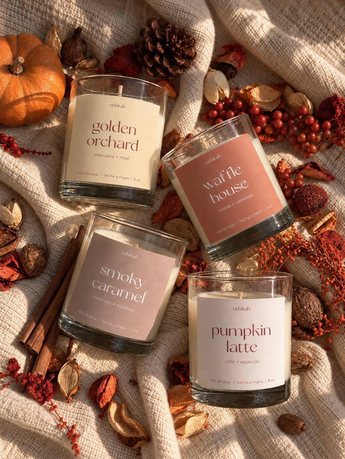 Four autumn-scented candles in glass jars surrounded by pumpkins and cinnamon sticks.