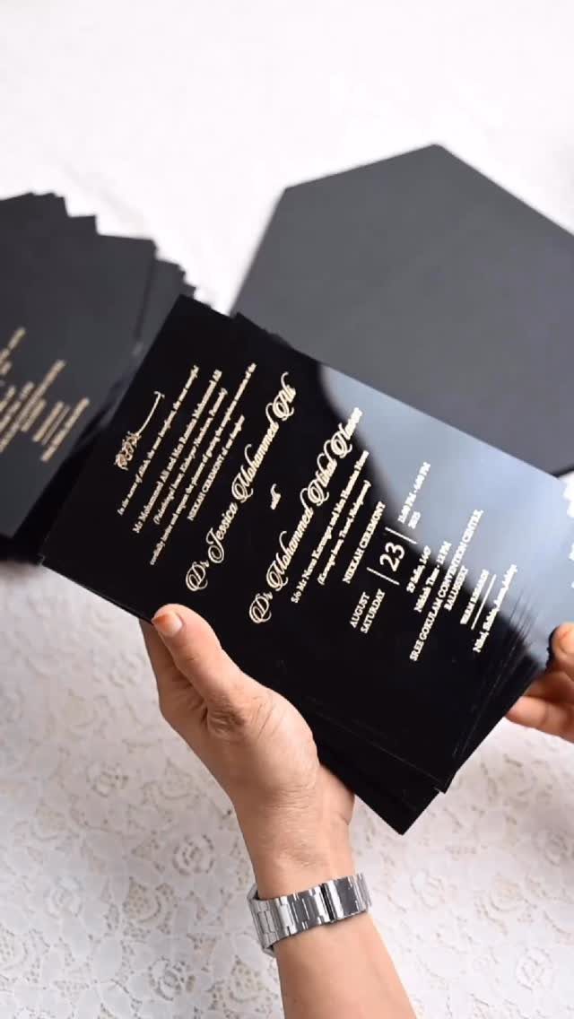 Black acrylic wedding invitations with gold foil lettering held by a person's hands.