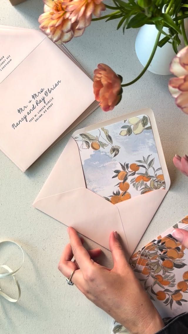 Pale pink envelopes with cursive calligraphy and citrus-patterned liners next to orange flowers.