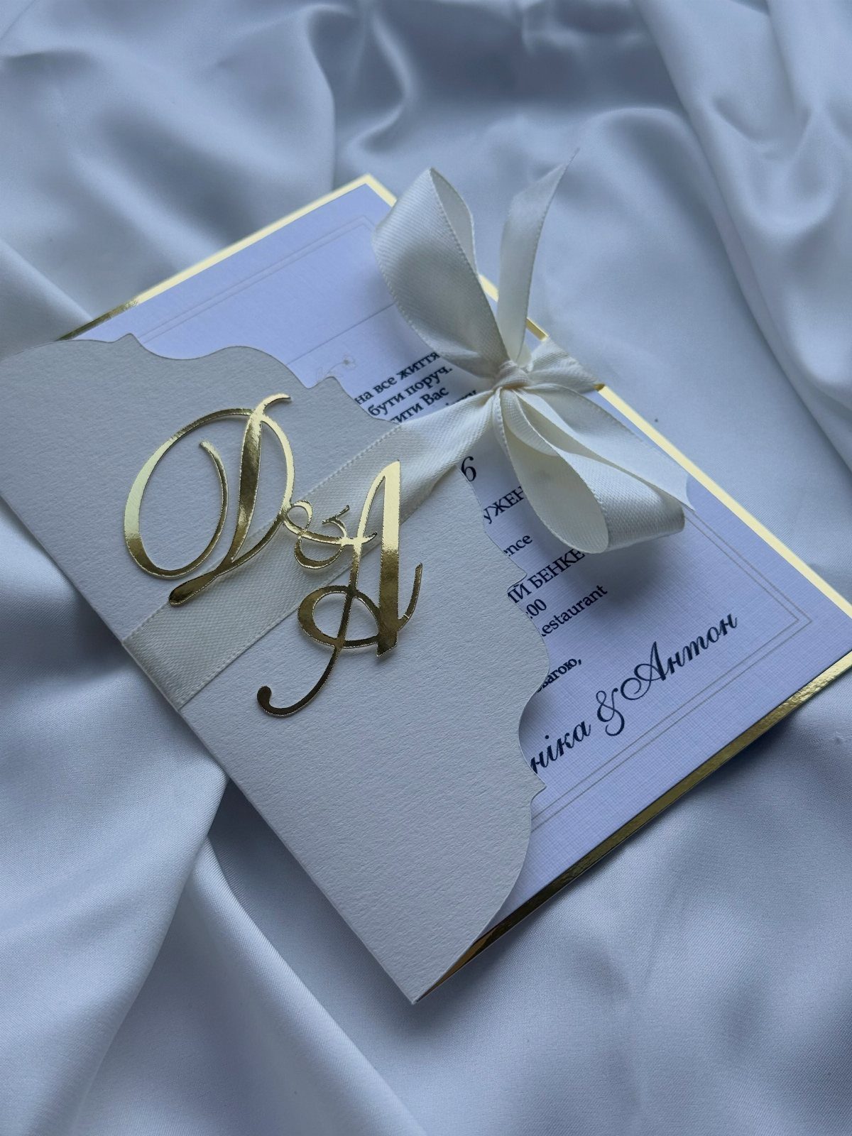 Formal wedding invitation with gold monogram and white satin bow on white silk fabric