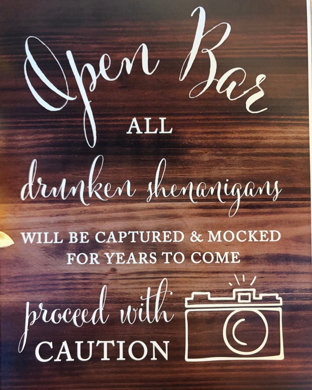 Wooden open bar sign with humorous warning about drunken shenanigans and a camera icon.