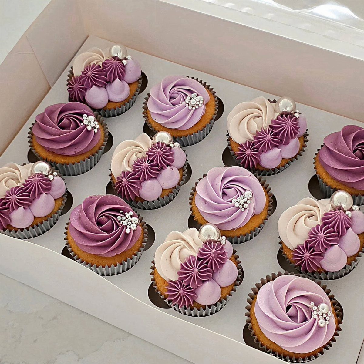 Cupcakes with purple and cream frosting and edible pearls in a white box