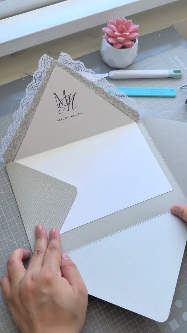 Wedding invitation suite with lace trim and monogram held by two hands.