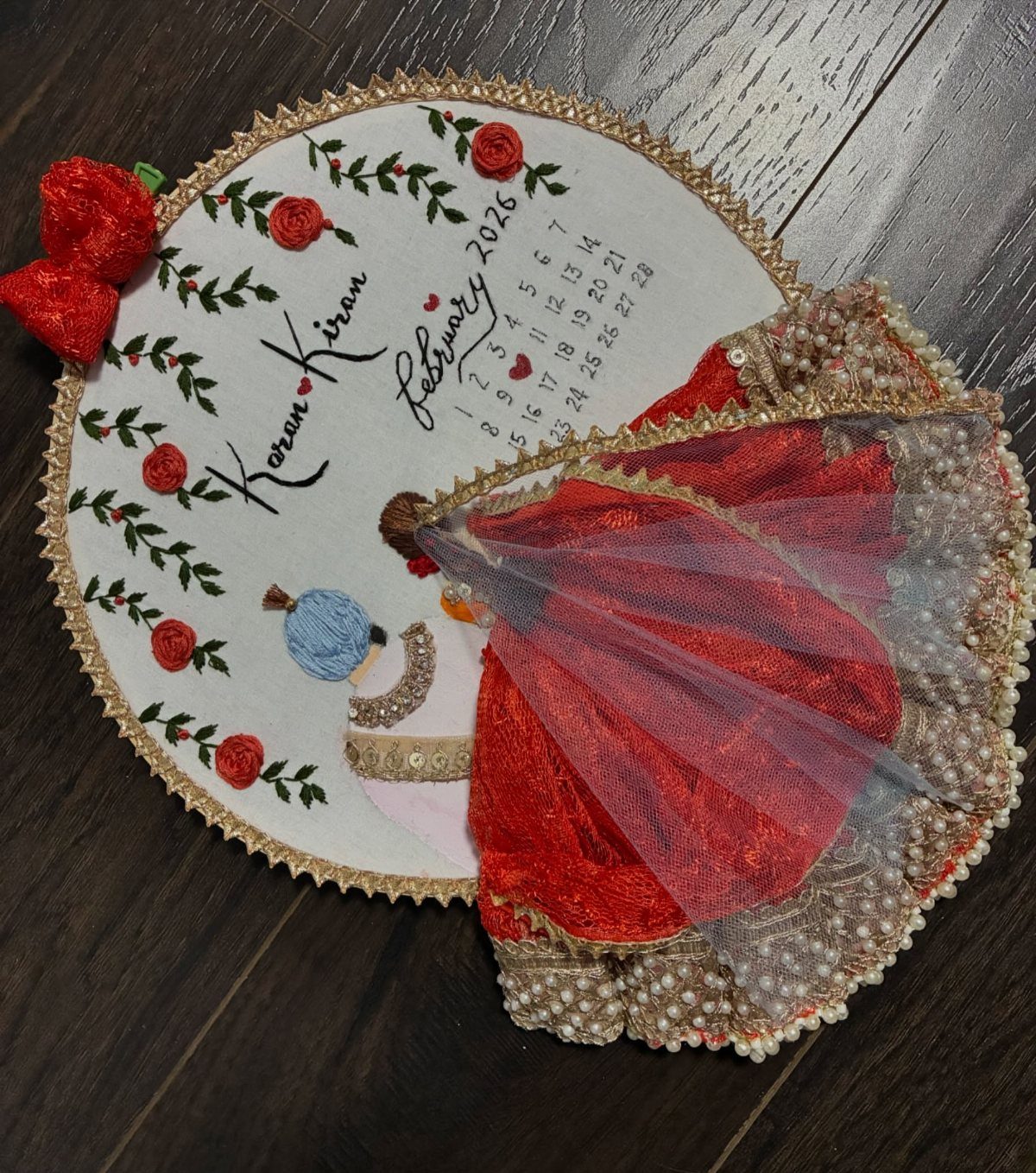 Embroidered oval wall art featuring a couple, a calendar, and red roses.