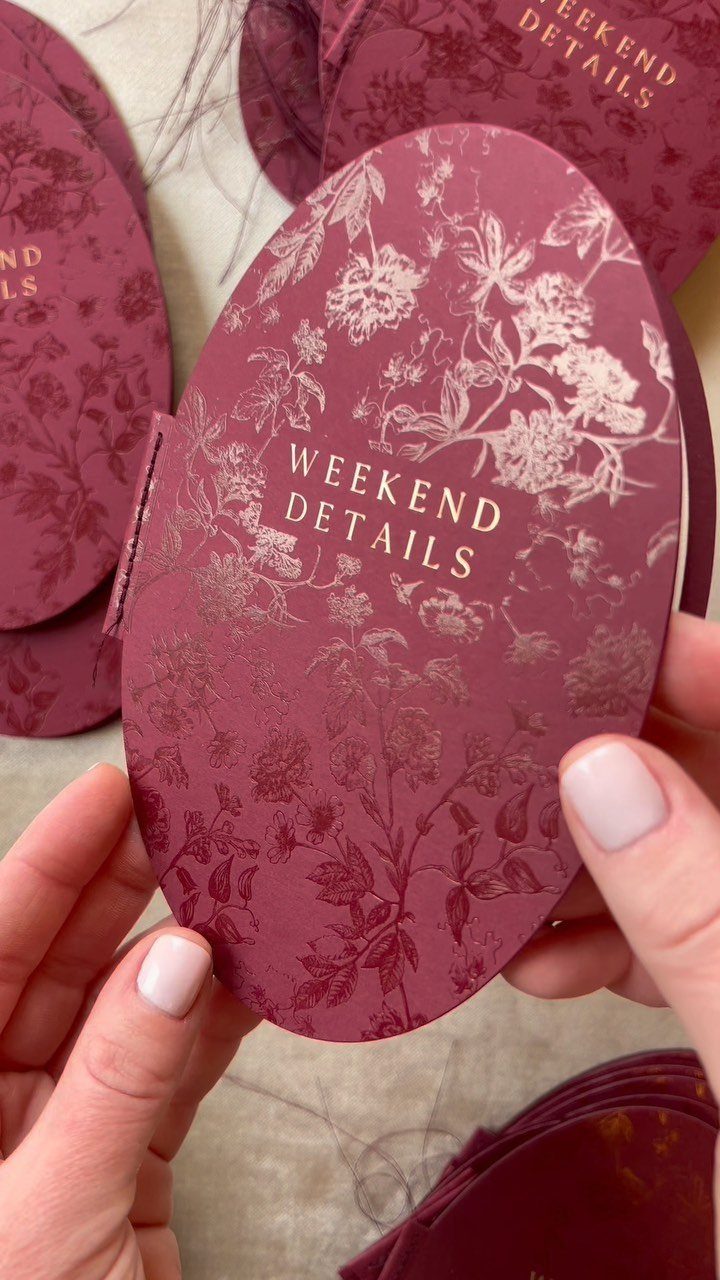 Burgundy oval wedding card with gold foil floral patterns and weekend details text.