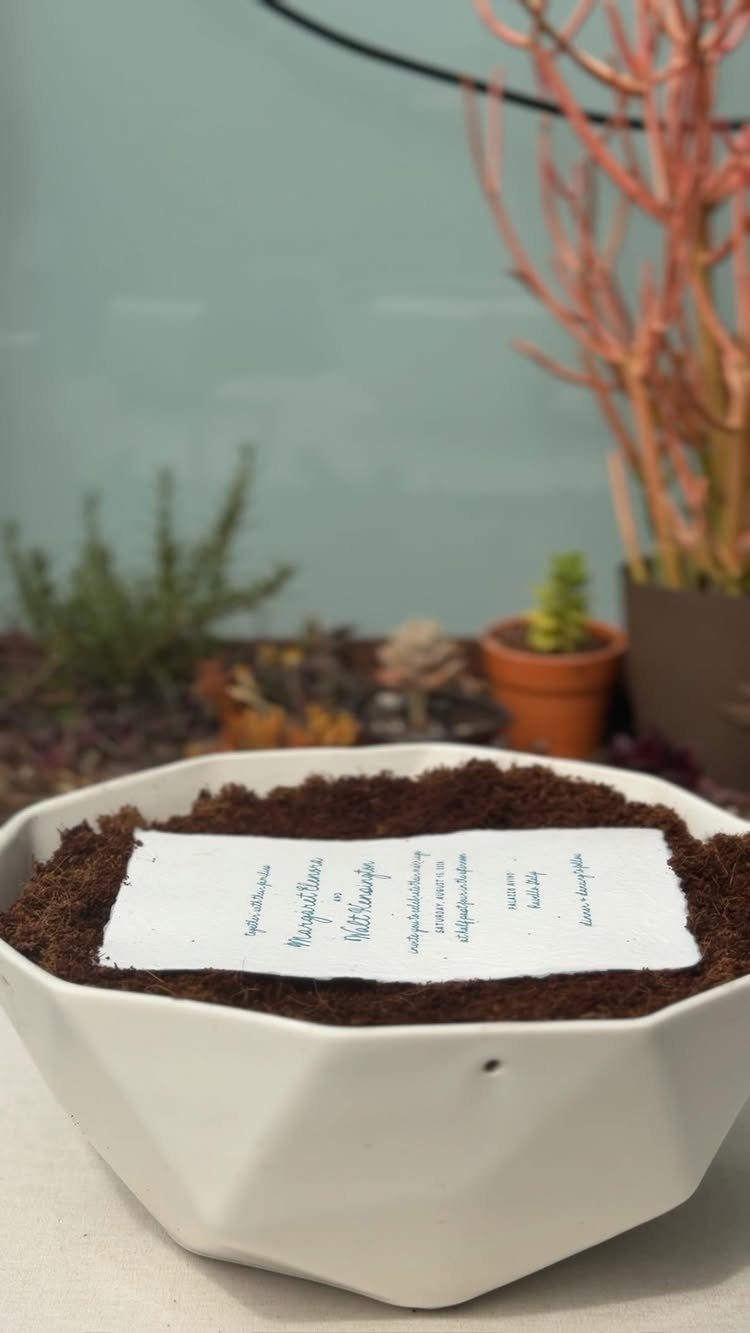 Handwritten invitation card resting on dark soil inside a white geometric ceramic bowl