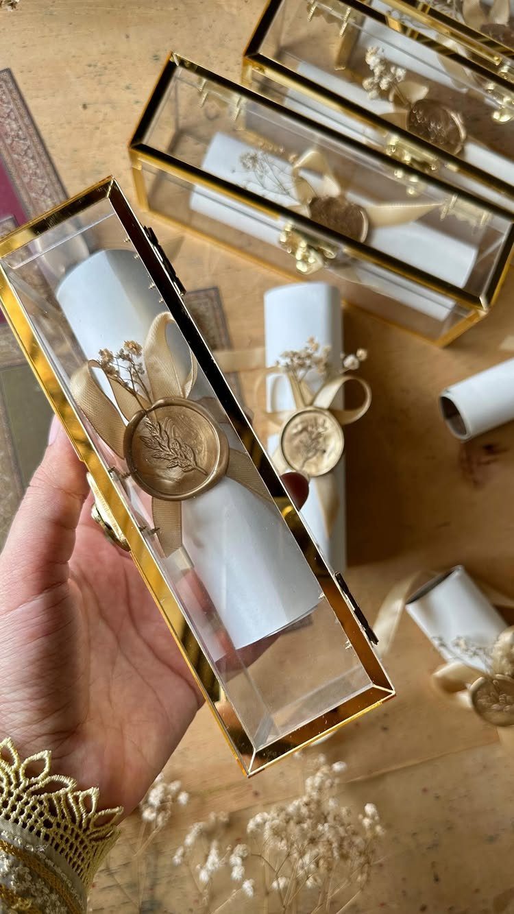 Rolled white wedding invitation with gold ribbon and wax seal in gold-edged box.