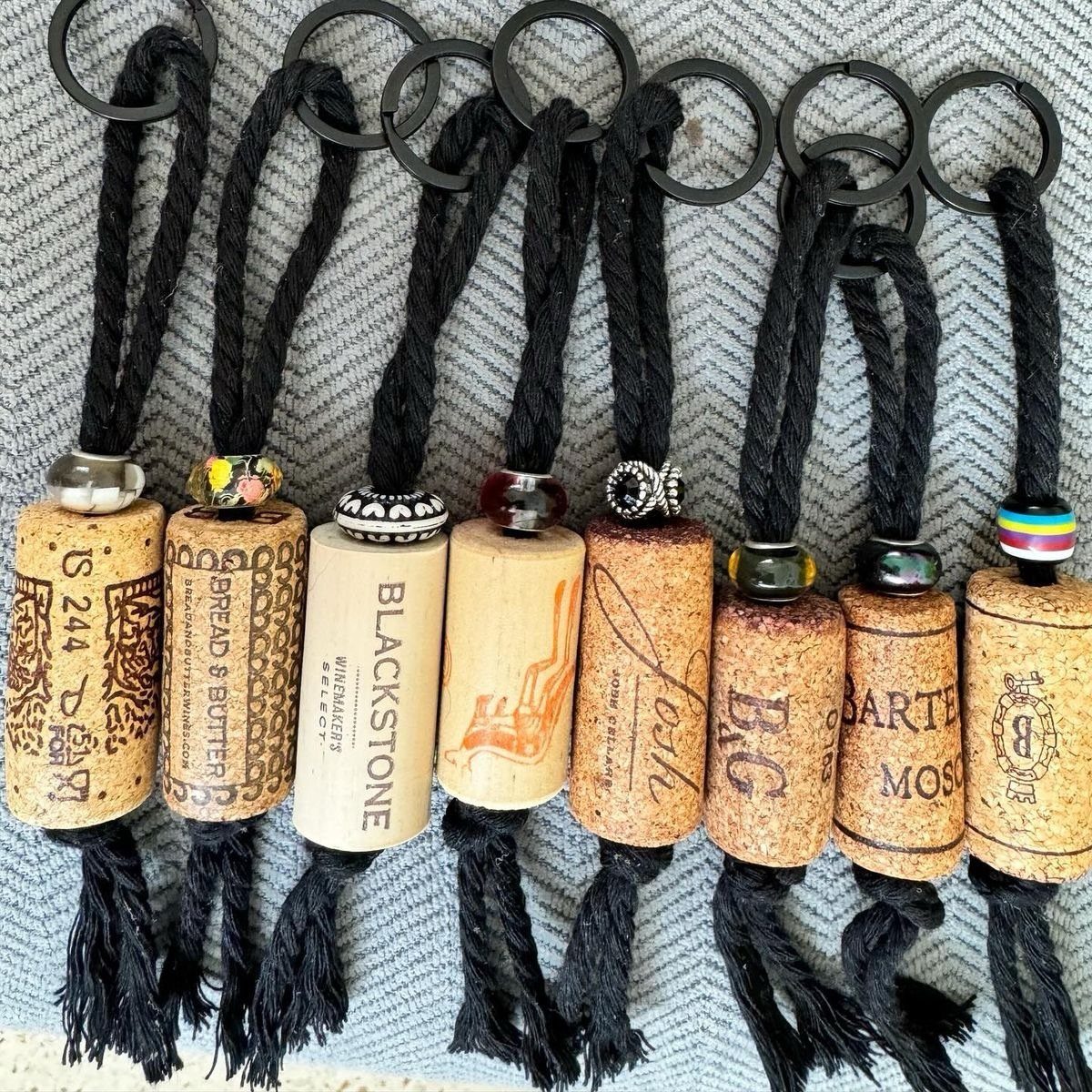 Wine cork keychains with black cords, decorative beads, and tassels on grey fabric.