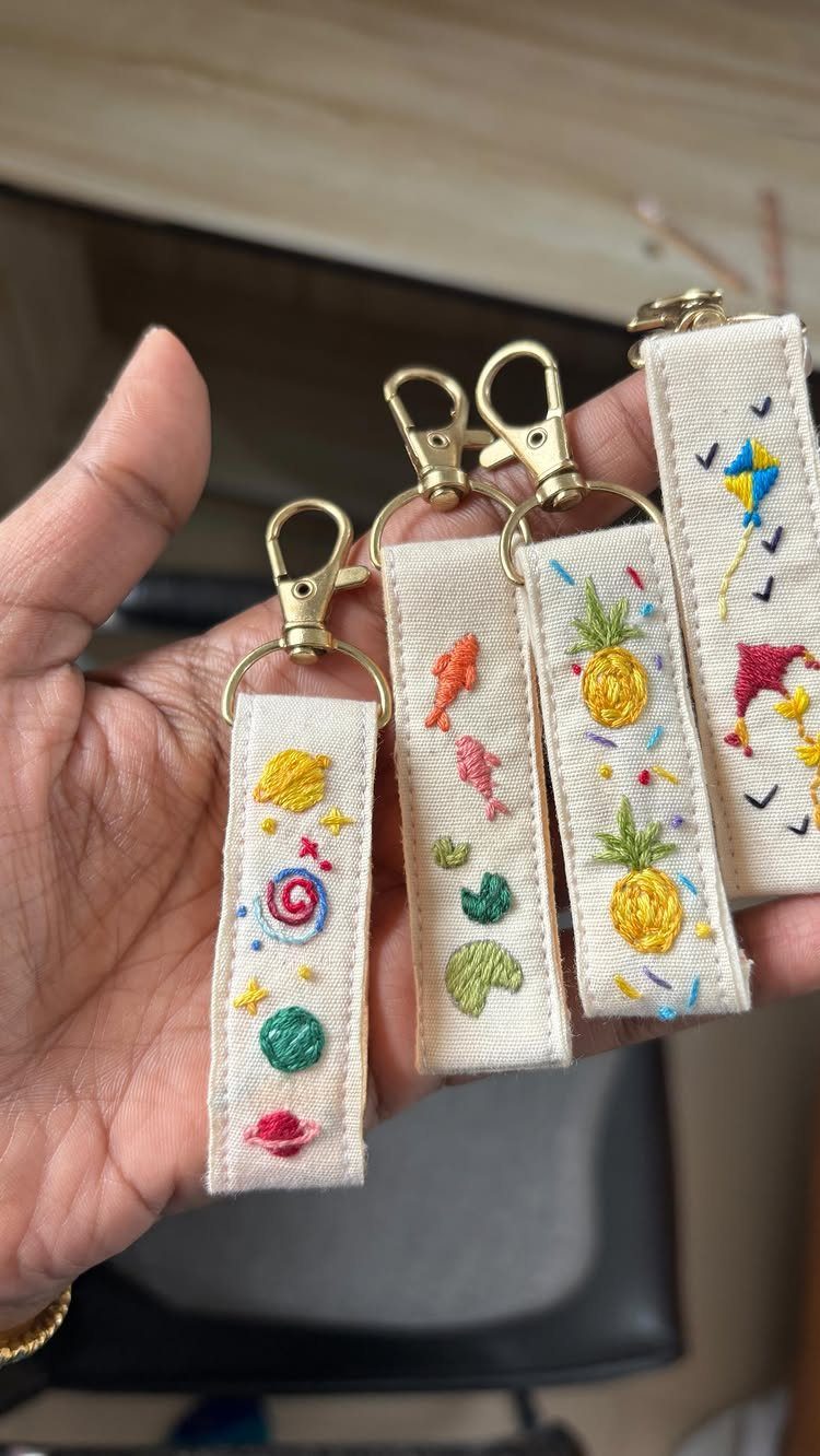 Embroidered fabric wristlet keychains with colorful designs held in a person's hand.