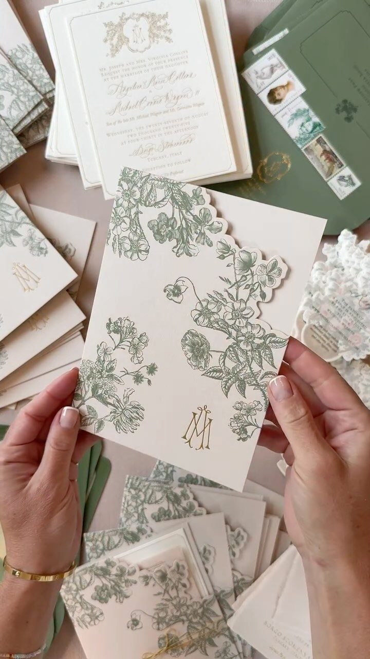 Hands holding a cream wedding invitation with green floral letterpress designs and gold monogram.