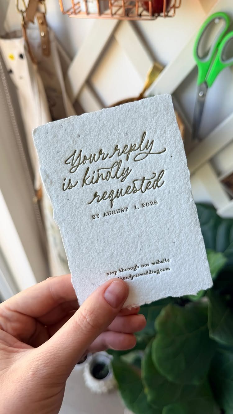 Textured white deckle-edged paper card with gold letterpress calligraphy held by a hand.