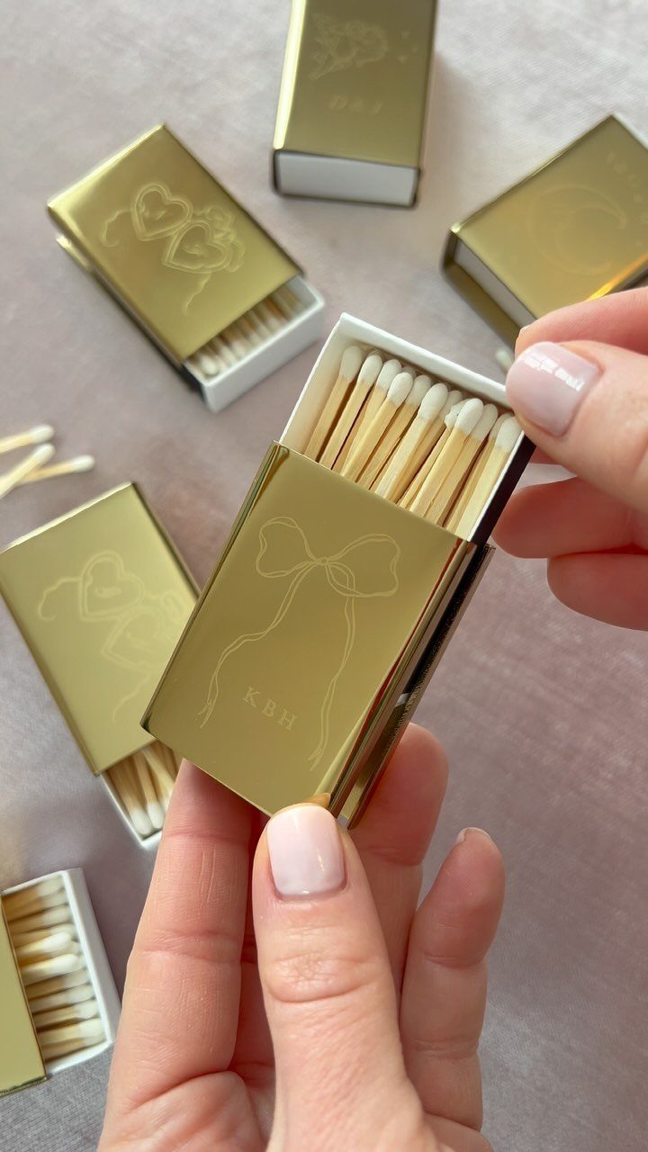 Gold metallic matchboxes with bow designs and initials on a pink background