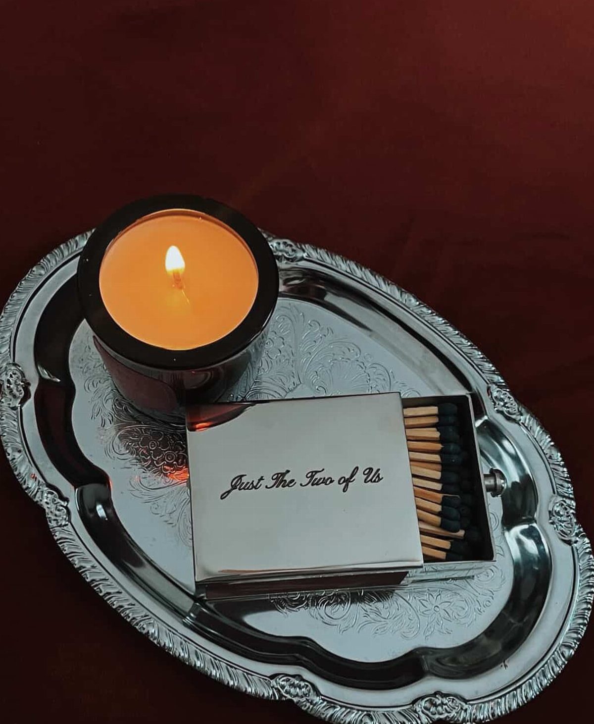 Lit candle and silver matchbox with text on an ornate silver tray.