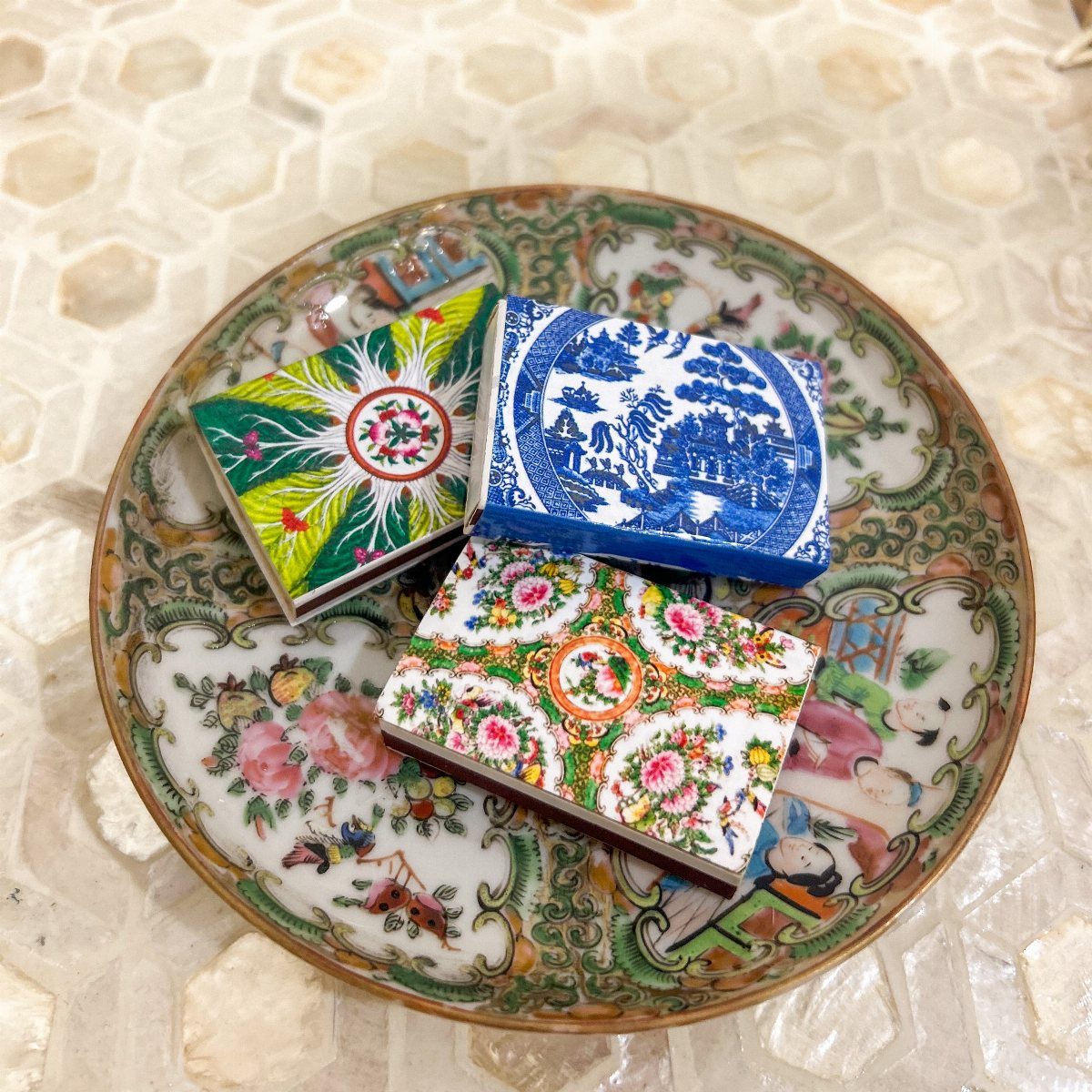 Three vintage-patterned matchboxes resting on a decorative ceramic plate with floral designs.
