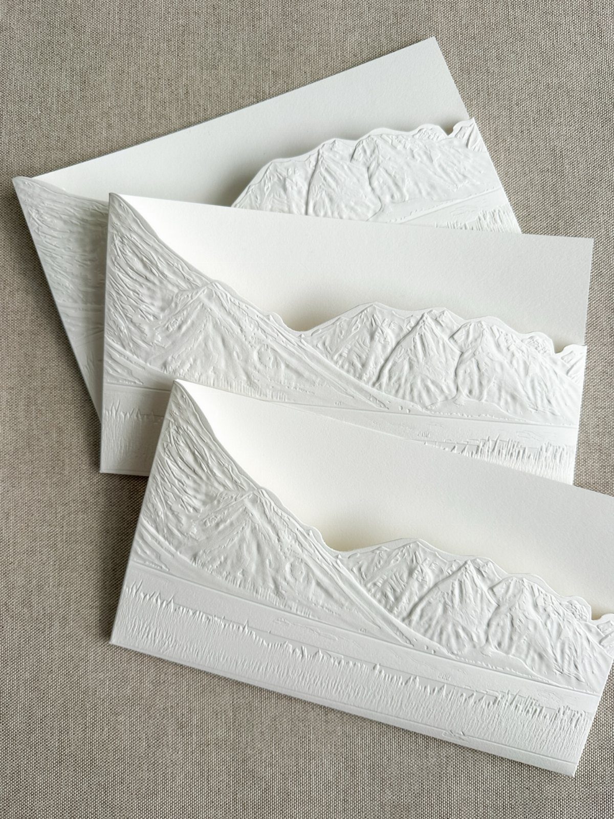 White embossed wedding invitations with detailed mountain range textures on linen fabric.